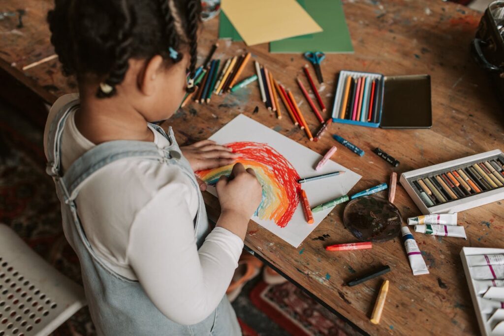 pexels photo 7025505 Young child creating a vibrant rainbow drawing at a wooden table with various art supplies.