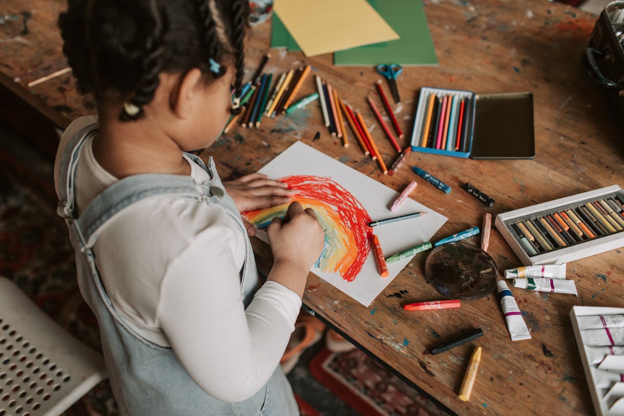 gallery-5 Young child creating a vibrant rainbow drawing at a wooden table with various art supplies.