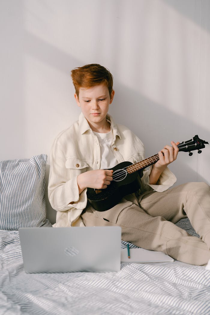 gallery-1 Redheaded preteen boy in beige outfit playing a ukulele while learning from a laptop at home.