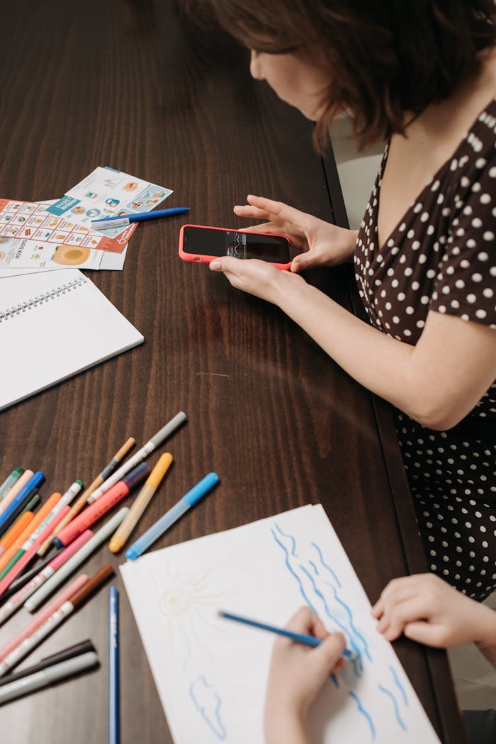 about-01 A mother supervises her child drawing at a table while holding a smartphone in a cozy indoor setting.